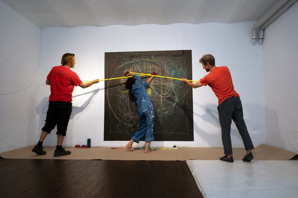 Anna Ruth, No strings attached, 2022. Three hours interactive performance, RAW – Relational Art Week, August 2022, Myymälä2, Helsinki. Materials: one artist, two retractable poles, tape, a wall, and pastels. Photo: Kamila Śladowska.